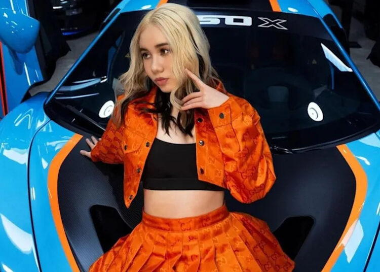 A woman with blonde hair wearing an orange outfit poses in front of a blue and orange sports car.