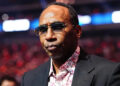 Stephen A. Smith wearing sunglasses and a patterned shirt under a dark blazer, standing in a venue with blurred lights in the background.