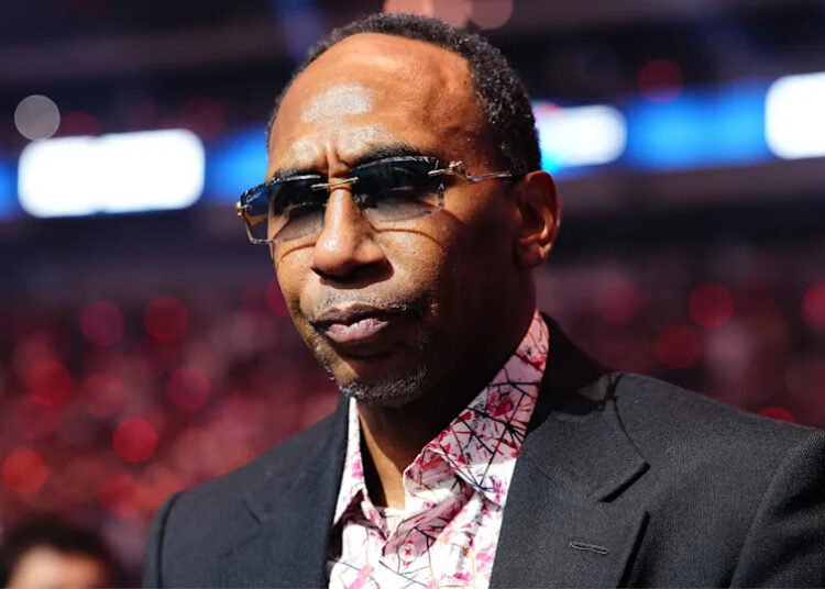 Stephen A. Smith wearing sunglasses and a patterned shirt under a dark blazer, standing in a venue with blurred lights in the background.