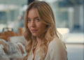 Sydney Sweeney standing in a coffee shop almost smiling in Anyone But You.