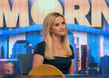 Reese Witherspoon as Bradley Jackson smiling at something off camera and sitting in front of The Morning Show sign in The Morning Show season 4.