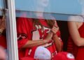 Taylor Swift watches Travis Kelce’s Kansas City Chiefs game from a suite, wearing a red team shirt, photographed through a glass barrier.
