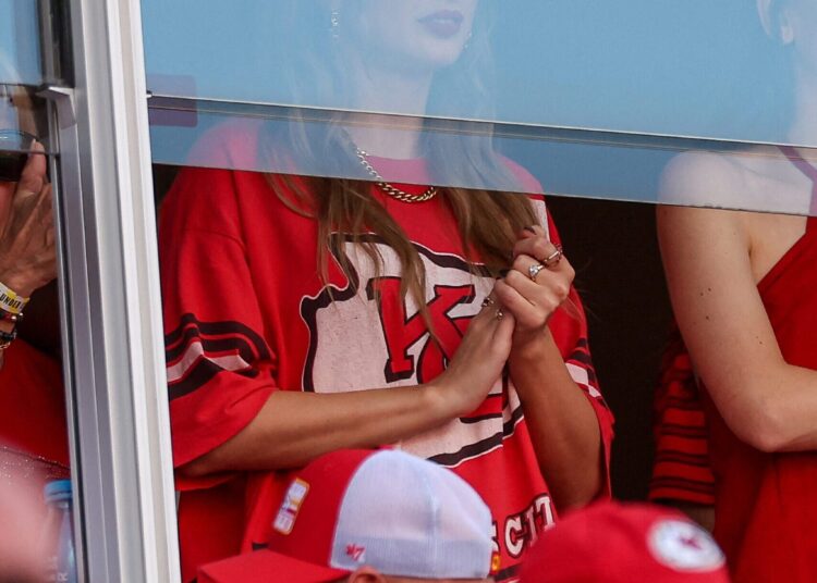 Taylor Swift watches Travis Kelce’s Kansas City Chiefs game from a suite, wearing a red team shirt, photographed through a glass barrier.