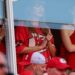 Taylor Swift watches Travis Kelce’s Kansas City Chiefs game from a suite, wearing a red team shirt, photographed through a glass barrier.