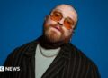 The image shows a bearded individual with tattoos on their head, wearing orange-tinted glasses, a black striped jacket over a light-coloured shirt, and a chain necklace. The background is a solid blue color.