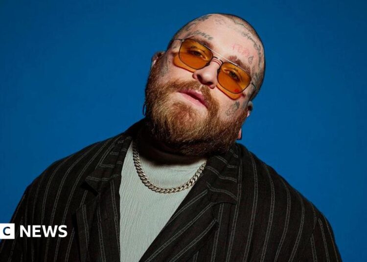 The image shows a bearded individual with tattoos on their head, wearing orange-tinted glasses, a black striped jacket over a light-coloured shirt, and a chain necklace. The background is a solid blue color.