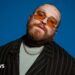 The image shows a bearded individual with tattoos on their head, wearing orange-tinted glasses, a black striped jacket over a light-coloured shirt, and a chain necklace. The background is a solid blue color.