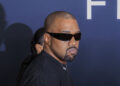 Kanye West wearing black sunglasses and a black shirt looks to the side while standing against a dark blue background.