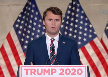 Charlie Kirk in a navy blue suit and striped tie speaks at a podium with “Trump 2020” signage, standing in front of American flags.
