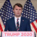 Charlie Kirk in a navy blue suit and striped tie speaks at a podium with “Trump 2020” signage, standing in front of American flags.