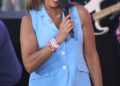 Hoda Kotb speaks into a microphone while wearing a light blue sleeveless blazer and white pants at an outdoor event.