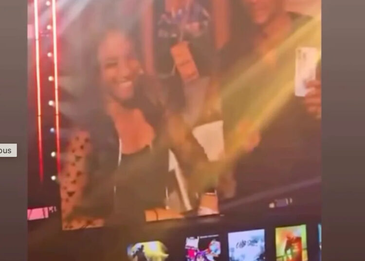 A large screen shows a woman smiling and waving at an event. The screen is surrounded by bright lights and a crowd below.