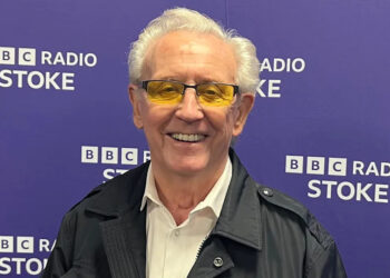 Tony Christie, with short white hair, yellow tinted glasses, a black jacket and a white shirt, smiles while he stands in front of a purple background that says BBC Radio Stoke in white letters.