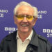 Tony Christie, with short white hair, yellow tinted glasses, a black jacket and a white shirt, smiles while he stands in front of a purple background that says BBC Radio Stoke in white letters.