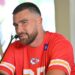 Travis Kelce speaks at a press conference wearing a red Kansas City Chiefs shirt with the team logo.