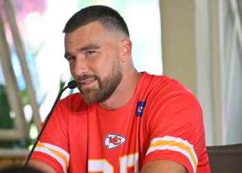 Travis Kelce speaks into a microphone while wearing a red Kansas City Chiefs shirt during a press event.