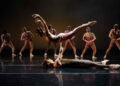 A dancer performs energetically among a group of fellow dancers, showcasing synchronized movements and vibrant expressions.