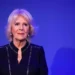 Blonde, short-haired woman with a serious expression dressed in black, standing in front of a blue background with a lit candle beside her
