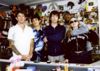 TURNSTILE poses for a polaroid picture in front of a shelf filled with records, knick knacks, and toys
