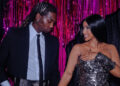 Offset and Cardi B stand together, smiling, in front of a pink and black backdrop. Offset is in a suit, and Cardi B wears a metallic dress.