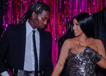 Offset and Cardi B stand together, smiling, in front of a pink and black backdrop. Offset is in a suit, and Cardi B wears a metallic dress.