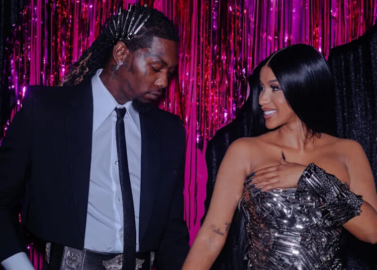 Offset and Cardi B stand together, smiling, in front of a pink and black backdrop. Offset is in a suit, and Cardi B wears a metallic dress.