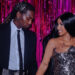 Offset and Cardi B stand together, smiling, in front of a pink and black backdrop. Offset is in a suit, and Cardi B wears a metallic dress.