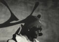 A person wearing sunglasses, a beanie, and a coat stands in front of a wall with abstract shadows. The image is in black and white.