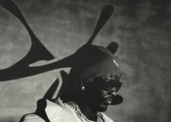 A person wearing sunglasses, a beanie, and a coat stands in front of a wall with abstract shadows. The image is in black and white.