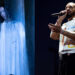 Julia Wolf stands behind a translucent blue curtain, and Drake stands with a microphone while performing on stage at night.