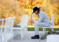 Jesse McBee in a grey suit and black hat, sitting among empty charis at his wedding and looking down at the ground with his hands folded
