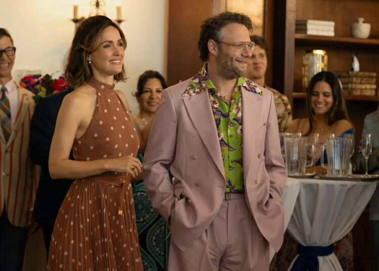 a woman in a polka dot dress stands next to a man in a pink suit at a large party