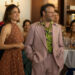 a woman in a polka dot dress stands next to a man in a pink suit at a large party