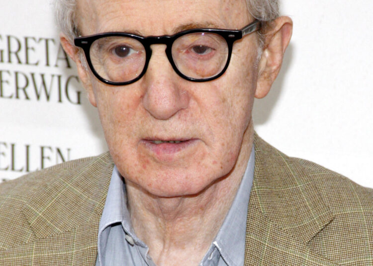 Woody Allen wearing glasses and a tan plaid blazer over a blue shirt at a public appearance.
