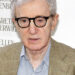 Woody Allen wearing glasses and a tan plaid blazer over a blue shirt at a public appearance.