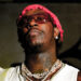 Young Thug wearing a red bandana, sunglasses, and multiple necklaces, looking upward with a serious expression.