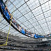 Interior of a large stadium with a transparent roof, a massive oval video screen displaying graphics, and a crowd of spectators.