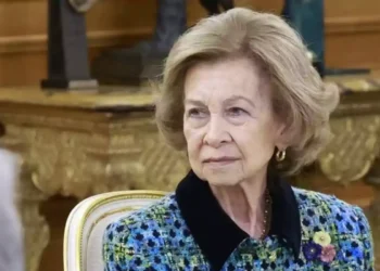 An older woman with blonde hair and elegant hairstyle wears a blue and green colored coat with a flower brooch while sitting on a chair against a background of decorative furniture.