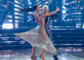 Corey Feldman and Jenna Johnson perform a ballroom dance under stage lights, with her sparkling silver dress flowing mid-spin.