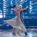 Corey Feldman and Jenna Johnson perform a ballroom dance under stage lights, with her sparkling silver dress flowing mid-spin.