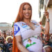 Cardi B, smiling and holding a phone, in a crowd wearing a colorful dress.