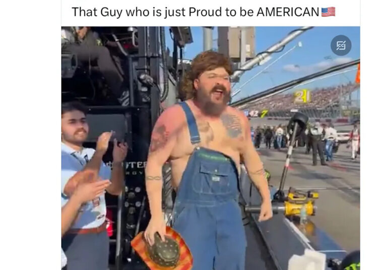 A man in overalls with tattoos and a mullet stands at a racetrack, holding a hat. Others nearby are clapping and smiling.