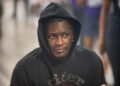 A person wearing a black hoodie with a pensive expression in a blurred background setting.