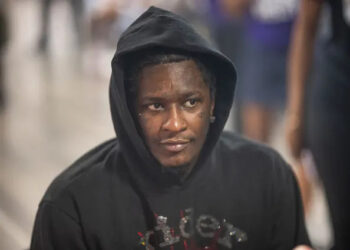 A person wearing a black hoodie with a pensive expression in a blurred background setting.