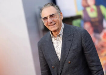 Michael Richards in a dark suit and round glasses smiles on the carpet