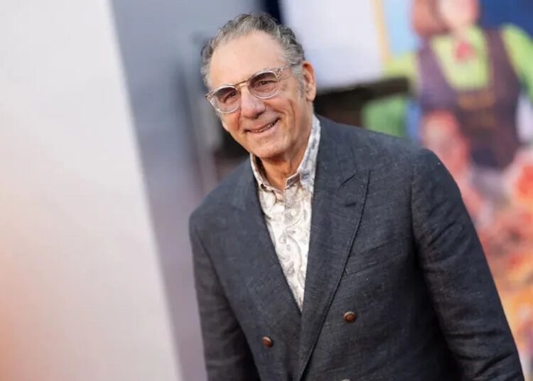 Michael Richards in a dark suit and round glasses smiles on the carpet