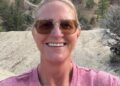 Christine Brown takes a selfie outdoors in front of rocky hills and pine trees, wearing sunglasses and a pink shirt.