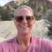Christine Brown takes a selfie outdoors in front of rocky hills and pine trees, wearing sunglasses and a pink shirt.