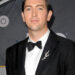 Nicholas Braun wears a black tuxedo with a bow tie and a silver branch-shaped pin on the lapel while posing at a formal event.