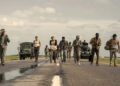 A group of people walking down a rural road, with military trucks and tanks in the background under a cloudy sky.